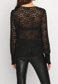 Haut en dentelle noire avec motifs floraux, manches longues et design semi-transparent. Le dos est transparent avec un col rond.