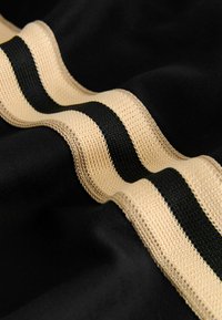 Schwarzer Stoff mit einem gewellten Muster, das zwei horizontale Streifen zeigt: einen strukturierten beigen Streifen und einen glatten schwarzen Streifen dazwischen.