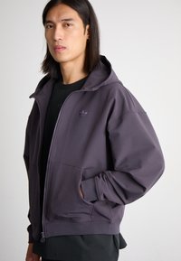 Zippered hooded jacket in dark purple fabric, featuring a front pocket and a subtle logo on the chest. Smooth texture, relaxed fit.