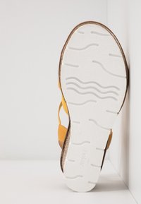 Semelle blanche d'une sandale jaune présentant des motifs ondulés et droits, placée à la verticale contre un mur d'angle blanc.