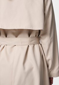 Nærbilde av baksiden på en beige trenchcoat med et belte knyttet i midjen og folder i stoffet under lommeryggklaffen.