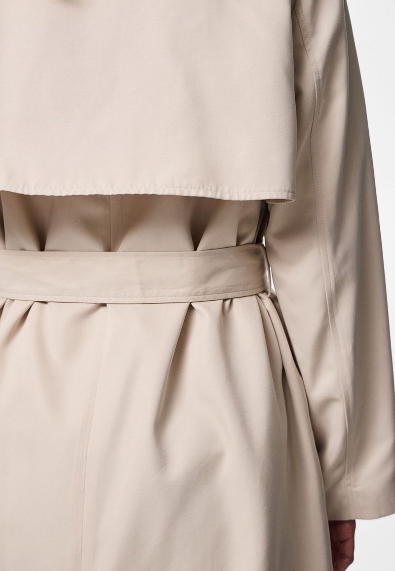 Nahaufnahme eines beigen Trenchcoats von hinten mit einem gebundenen Taillengürtel und plissierten Stoffdetails unter dem Rückenpatt.