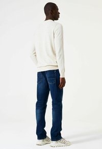 Man in een witte trui, donkerblauwe jeans en witte sneakers, staand en zijwaarts kijkend tegen een eenvoudige witte achtergrond.
