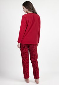 Mujer de pie descalza, de espaldas, vistiendo una blusa roja de manga larga y pantalones rojos con lunares negros.