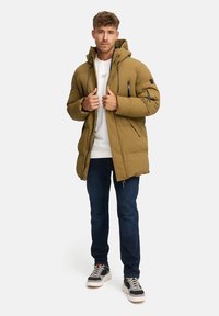 Gevoerde khaki parka met een capuchon, sluiting aan de voorkant en zijzakken, gedragen over een wit sweatshirt en een donkere spijkerbroek, gecombineerd met grijze sneakers.