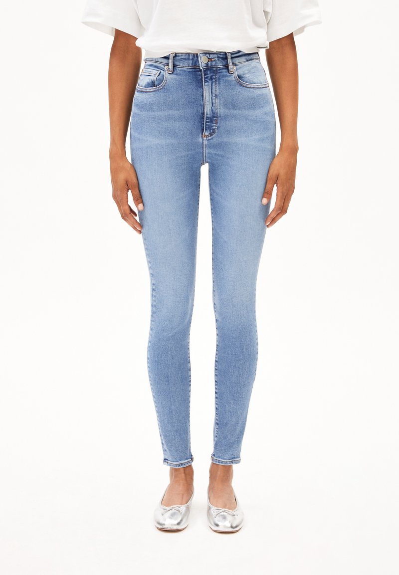 Jean slim taille haute bleu clair porté avec un haut blanc et des ballerines argentées, visible de la moitié du torse jusqu'aux pieds sur fond blanc.