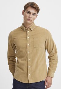 Bruine corduroy button-up blouse met een borstzak, lange mouwen, subtiele textuur en witte knopen. Model staat tegen een neutrale achtergrond.