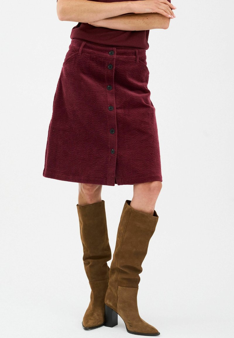 Bordo kordura suknja ravnog kroja, s prednjim kopčanjem na gumbi i bočnim džepovima. Nosena s smeškim zamšćenim čizmama do koljena.