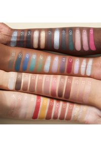 Quatre bras de teints différents montrant des échantillons de fards à paupières numérotés, dans des couleurs variées allant du nude aux teintes vives et métalliques.