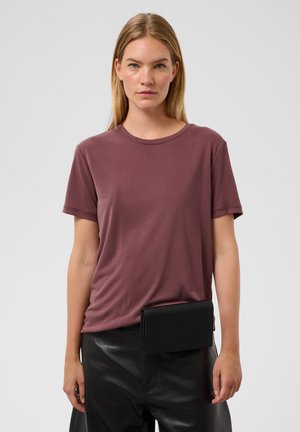 Femme aux cheveux blonds et raides portant un t-shirt mauve à manches courtes, un pantalon en cuir noir et un sac banane noir à la taille.