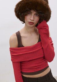 Rotes, geripptes Off-Shoulder-Oberteil, das über einem braunen Tanktop getragen wird, mit langen Ärmeln bis zu den Handgelenken. Ein brauner Pelzhut komplettiert den Look.