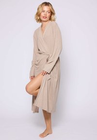 Beige wikkelrobe met lange mouwen, gemaakt van zachte stof. Heeft een strik in de taille, witte bies langs de halslijn en twee voorzakken.