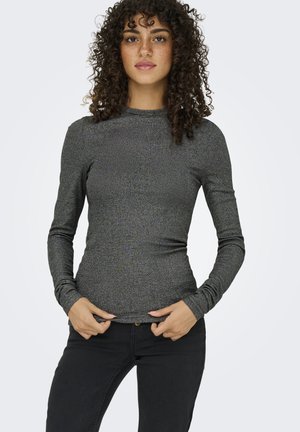 Donna con capelli ricci che indossa una maglia grigia aderente a maniche lunghe e pantaloni neri, tenendo l'orlo della maglia con entrambe le mani.