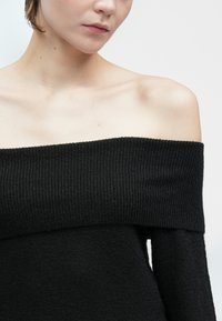 Kvinne iført en svart ribbestrikket genser med avdekkede skuldre, som viser kragebeina og en del av ansiktet mot en enkel lys bakgrunn.