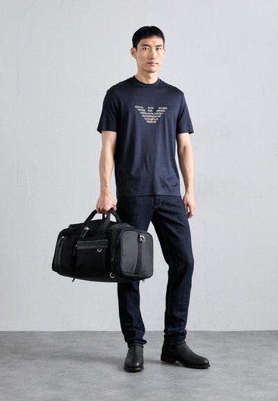 Navy t-shirt with a reflective logo, black duffel bag with multiple compartments, dark jeans, and black ankle boots in a neutral background.