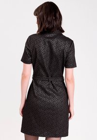 Woman with shoulder-length dark hair wearing short-sleeve, belted black dress with textured diamond pattern, standing facing away on plain background.