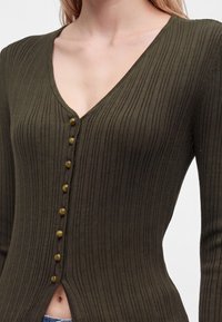 Gros plan d'une personne portant un cardigan ajusté, côtelé, vert olive, avec des boutons sur le devant et un col en V.