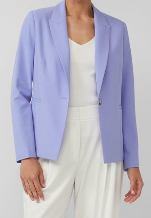 Vrouw draagt een lichtpaarse blazer over een wit V-hals topje en witte hoge taille broek, staat met handen ontspannen langs haar zij.