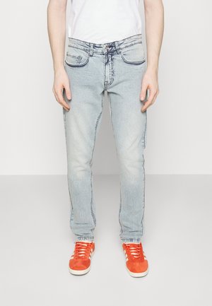 Persona con jeans desgastados de color azul claro y zapatillas deportivas de color naranja brillante, de pie frente a un fondo blanco liso.