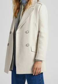 Beige wool peacoat with double-breasted buttons, notched lapels, textured details, and a side pocket. Worn over a checked shirt.