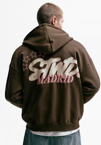 Brun hættetrøje med stort trykt logo med teksten "STWD" og "MADRID" i pink, omgivet af pink stjerner. Blødt stof, afslappet pasform.