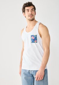 Canotta bianca con una tasca floreale colorata sul lato sinistro. Il design include foglie tropicali e fiori.