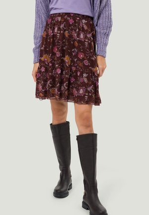 Frau, die einen lila Strickpullover, einen braunen Blumenrock und schwarze kniehohe Lederstiefel vor hellem Hintergrund trägt.