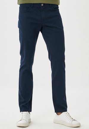 Mand iført marineblå slim-fit bukser og hvide sneakers, stående mod en ensfarvet hvid baggrund.