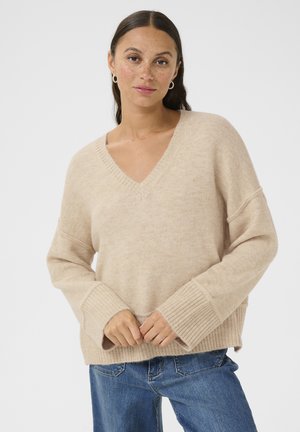Beiger, V-Ausschnitt-Pullover aus weichem Material, mit lockeren Ärmeln, gerippten Bündchen und einer entspannten Passform. Kombiniert mit blauen Jeans.