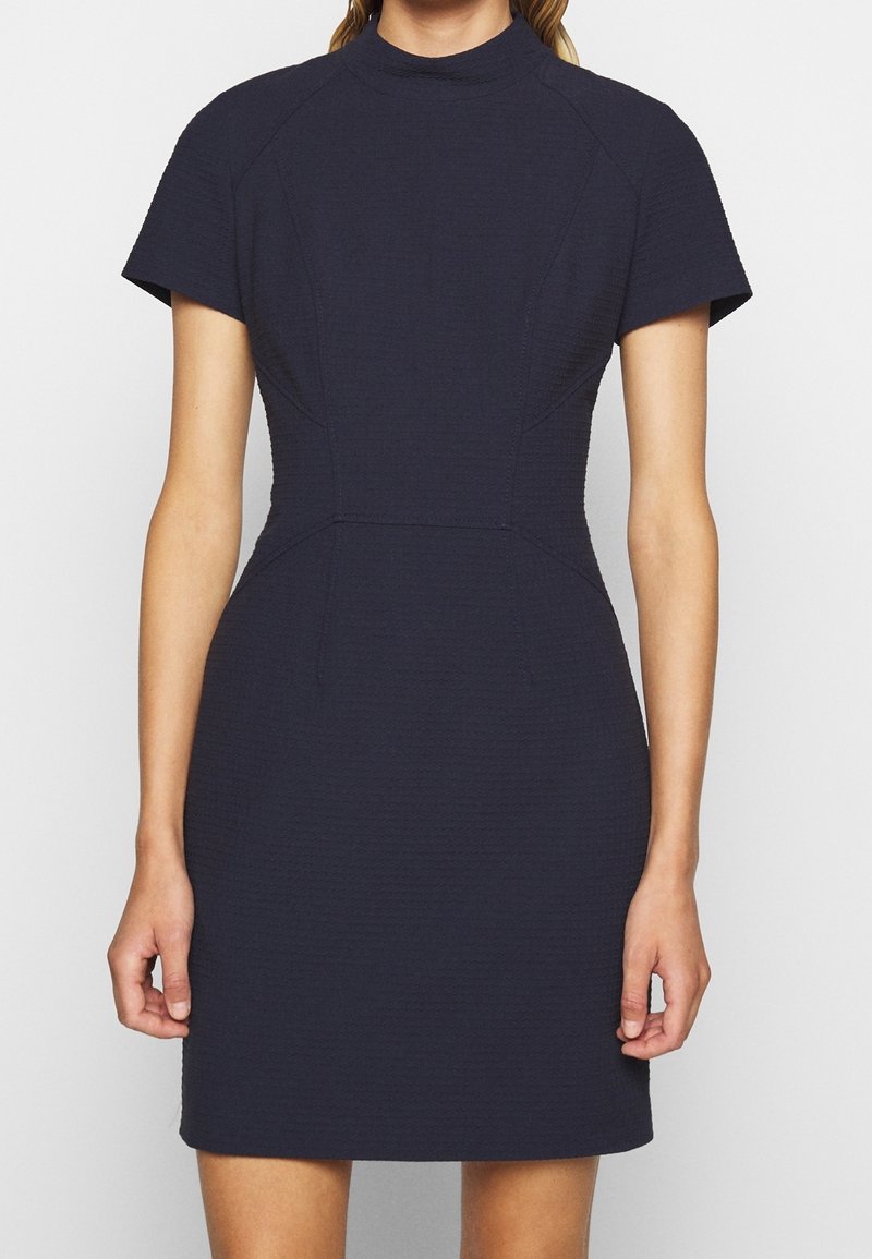 Robe bleu marine à manches courtes avec un col montant, un tissu texturé, une coupe ajustée, et des coutures décoratives le long du corsage et des manches.