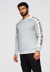 Sudadera gris con cuello redondo, mangas raglán y rayas blancas de marca en contraste. El material parece suave y casual.