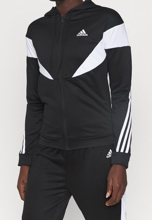 Czarna sportowa kurtka Adidas na zamek z białymi paskami na rękawach i piersi, noszona z dopasowanymi czarnymi spodniami z białymi paskami po bokach.