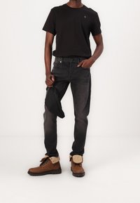 Camiseta negra, jeans gris oscuro y botas marrones con borde de pelo. Sosteniendo una pequeña bolsa negra. Atuendo simple y casual.