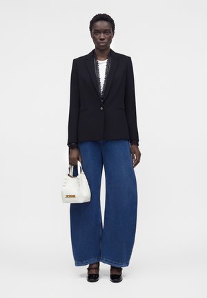 Donna con blazer nero sopra top bianco, jeans blu a gamba larga, scarpe nere, che tiene una piccola borsa bianca su sfondo semplice.