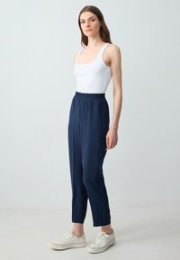 Eine Frau steht mit entspannten Händen da, tr�ägt ein weißes Tank-Top, marineblaue Hose mit elastischem Bund und weiße Turnschuhe vor einem schlichten, hellen Hintergrund.