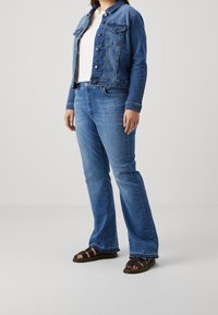 Veste en denim bleu avec deux poches avant, associée à un jean évasé bleu. Portée avec des sandales noires à lanières, présentant une coupe décontractée et un délavage clair.