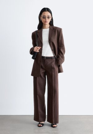 Femme portant un blazer marron oversize et un pantalon à jambes larges, chemise blanche, sandales noires, lunettes de soleil, et portant un sac à épaule noir.