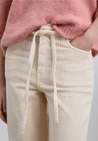 Pantalon en velours côtelé crème avec une taille à cordon, présenté aux côtés d'un pull tricoté rose doux, mettant en valeur la texture et les détails du tissu.