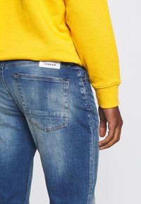 Denham blå denimjeans med blekning, femfickdesign och en vit etikett på baksidan. Kombinerat med en ljusgul sweatshirt.