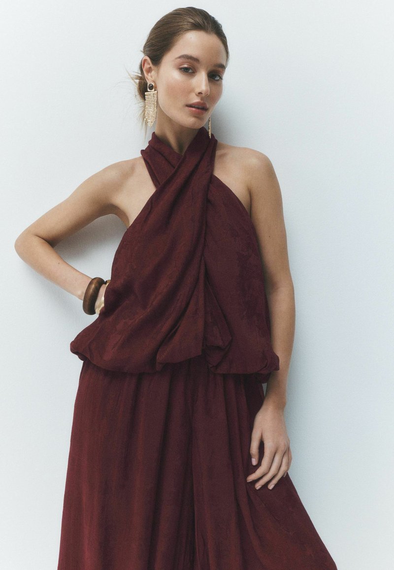 Mujer con top halter sin mangas rojo oscuro y falda, con grandes pendientes colgantes y una pulsera de madera, posando sobre fondo blanco.