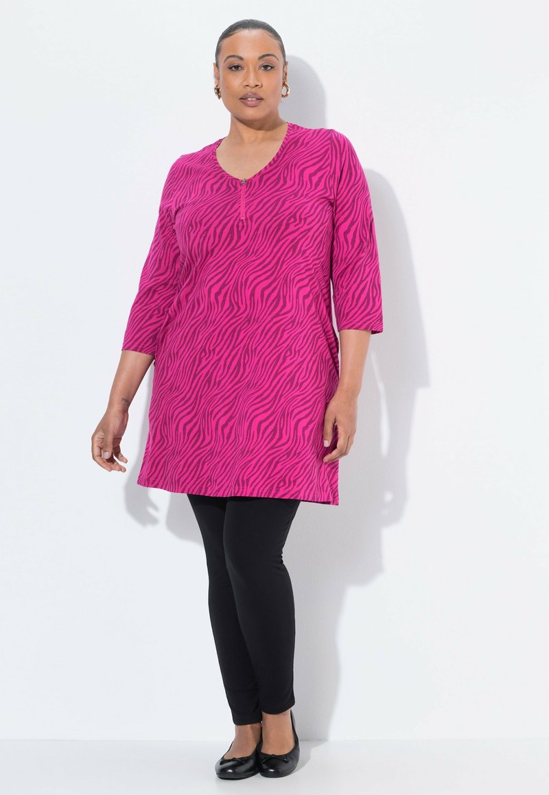 Rosa Tunika mit Zebramuster, mit V-Ausschnitt und 3/4-Ärmeln, kombiniert mit schwarzen Leggings und flachen Schuhen. Glatte Stofftextur.