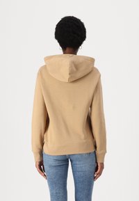 Haut à capuche kaki avec une poche frontale, des poignets et un ourlet côtelés ; tissu doux ; comporte une capuche avec cordon. Porté avec un jean en denim bleu.