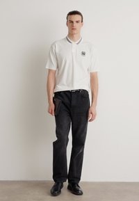 Witte polo met een grijsgestreepte kraag, geborduurd logo op de borst; gecombineerd met zwarte jeans en zwarte schoenen, houdt een zwarte portemonnee vast.