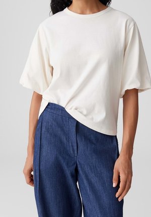 Personne portant une chemise blanche ample à manches courtes, partiellement rentrée dans un pantalon en jean bleu taille haute à jambes larges.