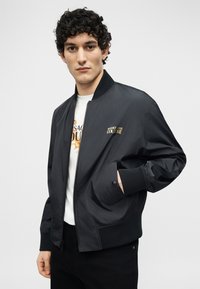 Νέος με καστανά σγουρά μαλλιά φοράει μαύρο μπουφάν Versace bomber και λευκό t-shirt με στάμπα, στέκεται μπροστά σε απλό φόντο.