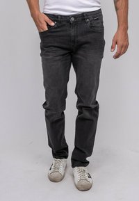 Jean en denim noir à coupe slim, avec des poches latérales, une fermeture à bouton et des décolorations subtiles. Associé à des baskets blanches.