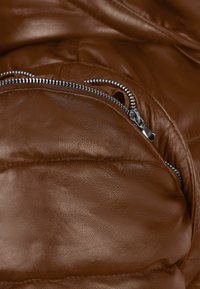 Giacca in pelle marrone con tasca con zip in metallo argento, texture liscia e sezioni di tessuto leggermente imbottite.