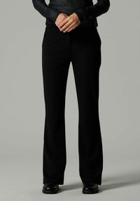 Isik, kellel on seljas mustad laiad püksid, mustad saapad ja tume pikakäised ülikond, hoiab käsi koos rinnal, seistes ühetoalise tausta ees.