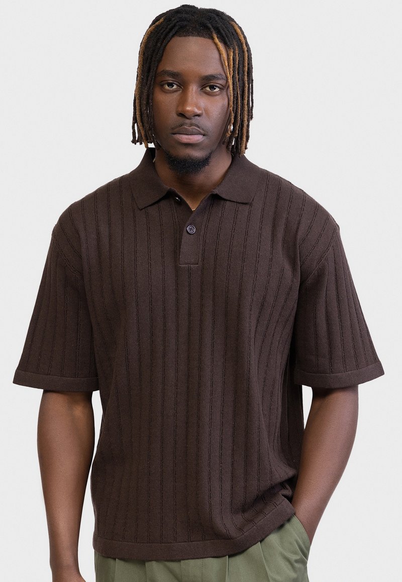 Homme aux dreadlocks mi-longs portant un polo en maille à manches courtes de couleur marron et un pantalon vert, debout devant un fond uni.