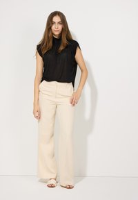 Schwarzes Kurzarmoberteil mit drapiertem Design, kombiniert mit hoch taillierten, cremefarbenen weiten Hosen und hellen Sandalen, vor einem weißen Hintergrund.